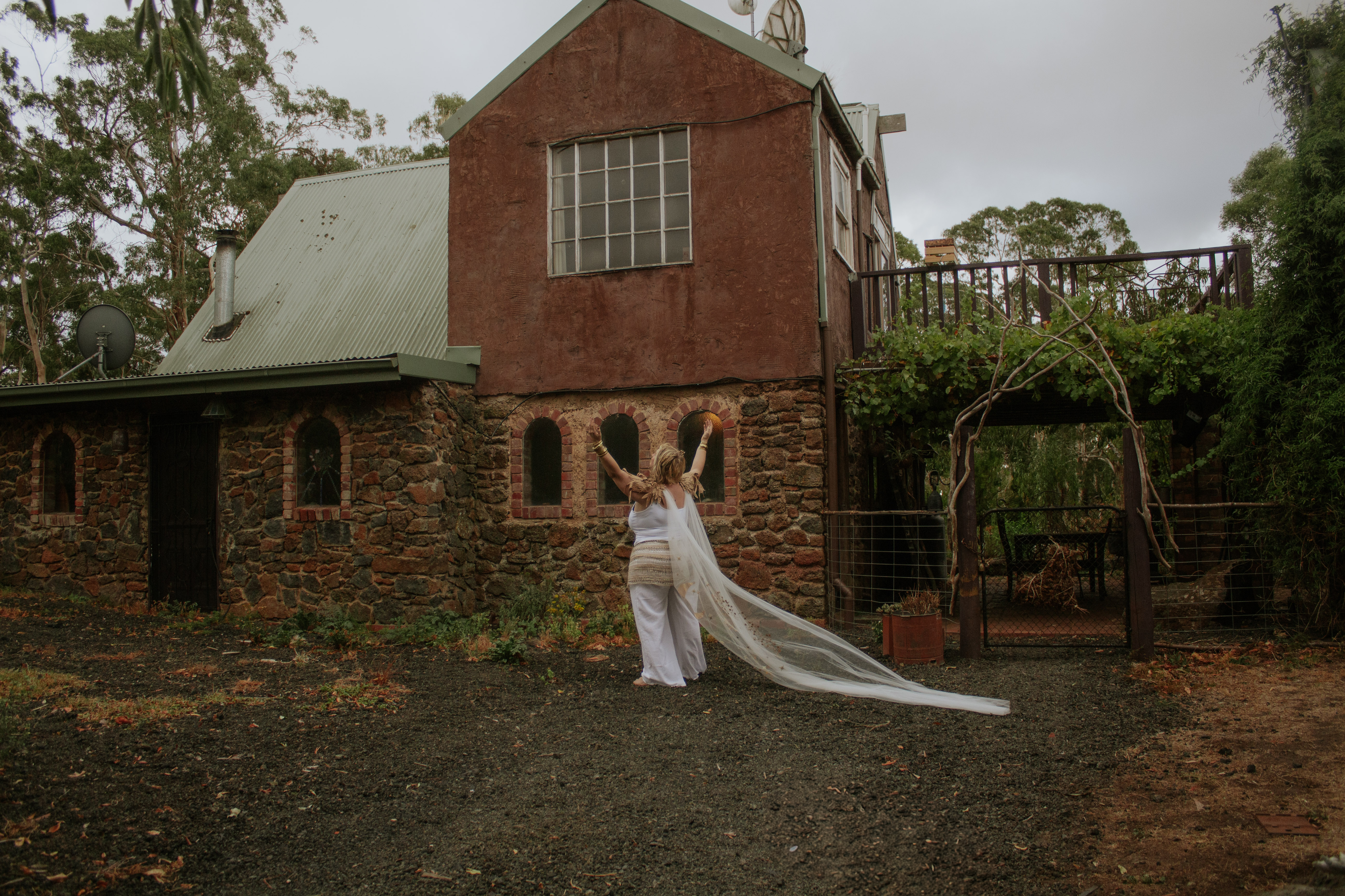 Woman in white with arms raised before the stone homestead, landscape