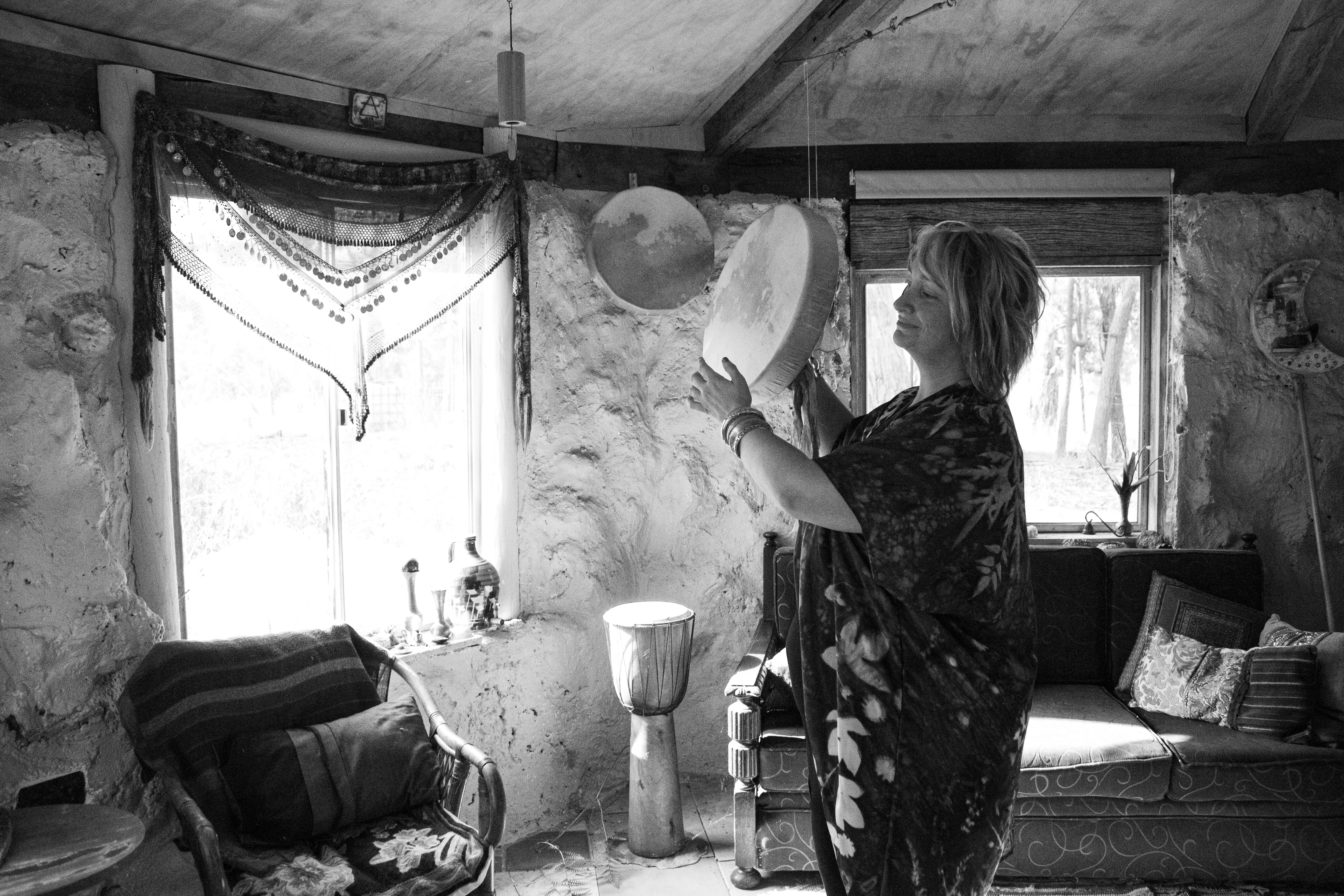 Black and white documentary photograph of a person in an artist's studio holding a drum, with natural light streaming through windows into a textured interior space