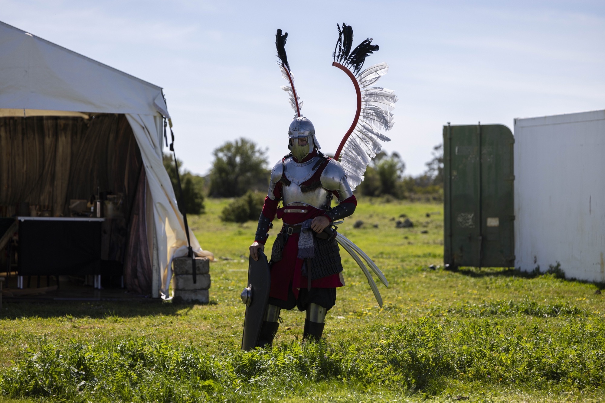 A Swordcraft player in elaborate silver and red armour with feathered wings on their helmet walks across a green field at the Pomborneit event