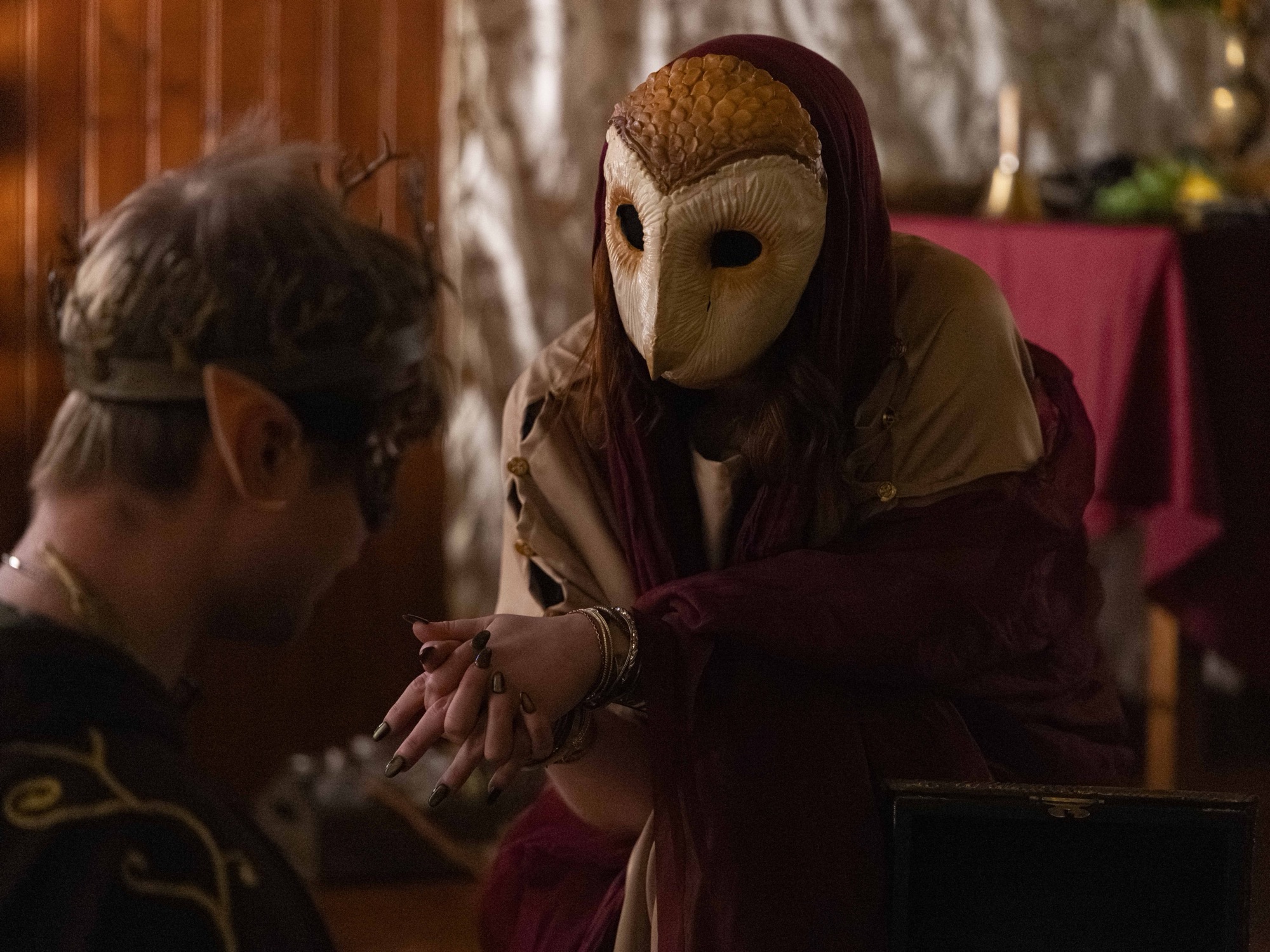 A Swordcraft player in an elaborate owl mask and robes leans in toward another costumed player in an intimate roleplay scene in a dimly lit interior