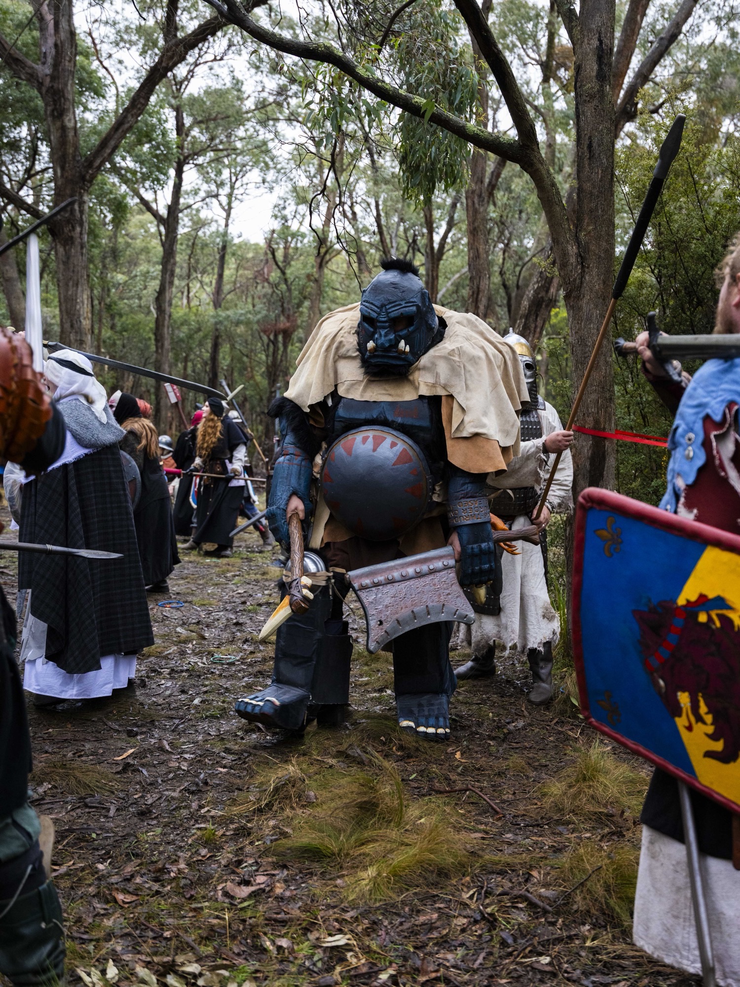 A player dressed as an enormous armoured orc creature with a foam hammer advances through a crowded battle scene among trees at Pomborneit