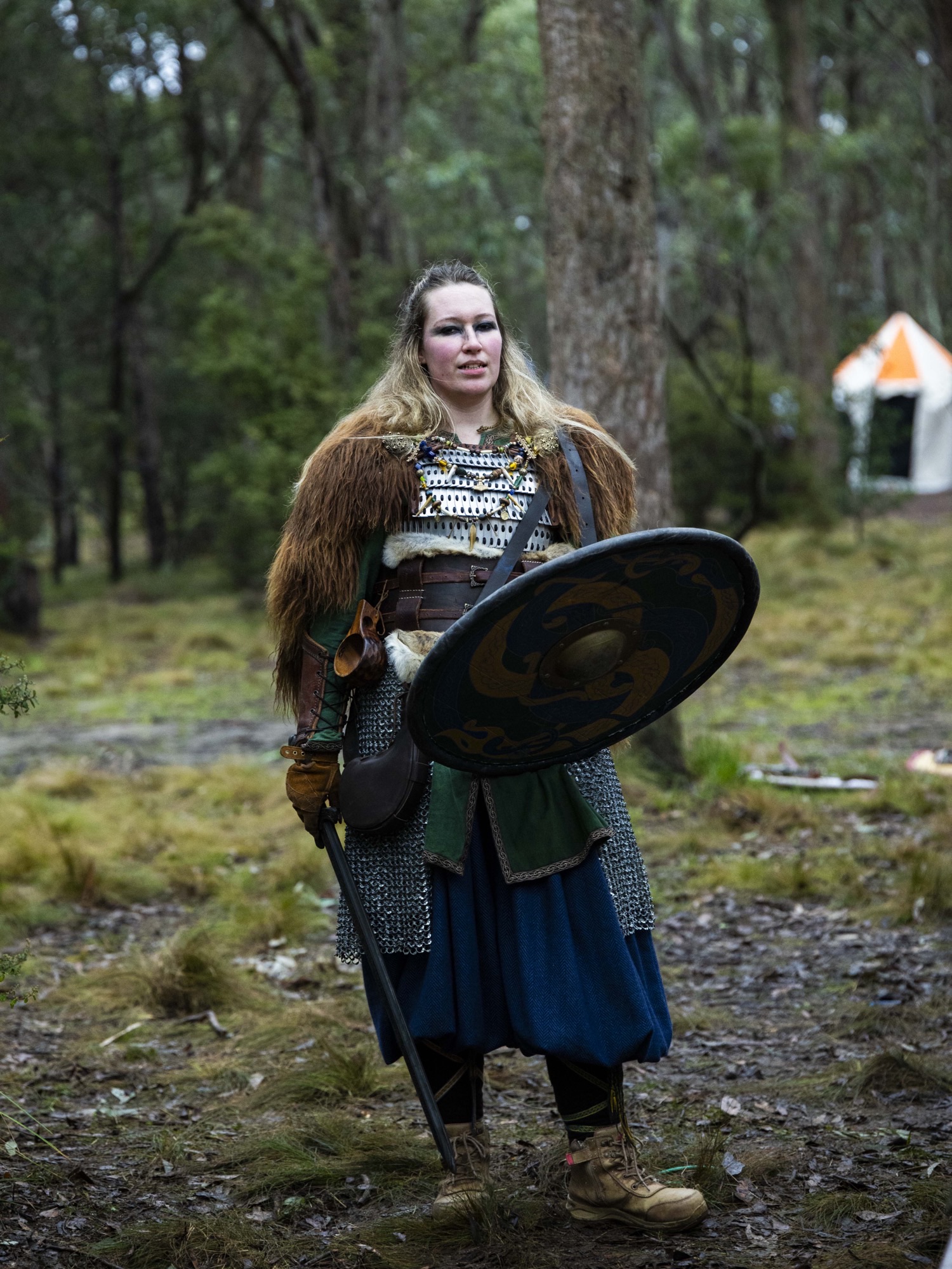 A female Swordcraft player in chainmail and fur armour holds a round shield decorated with a dragon, standing among trees at the Pomborneit event