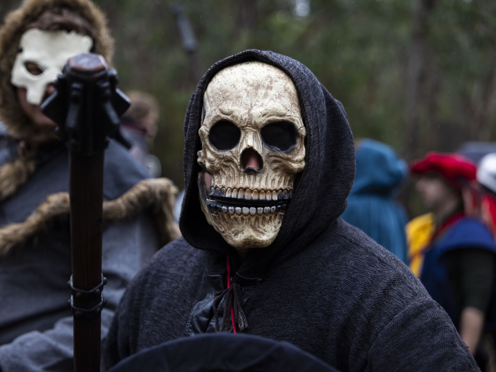 A masked undead Swordcraft player in a black hood stands among costumed players in the forest at Pomborneit