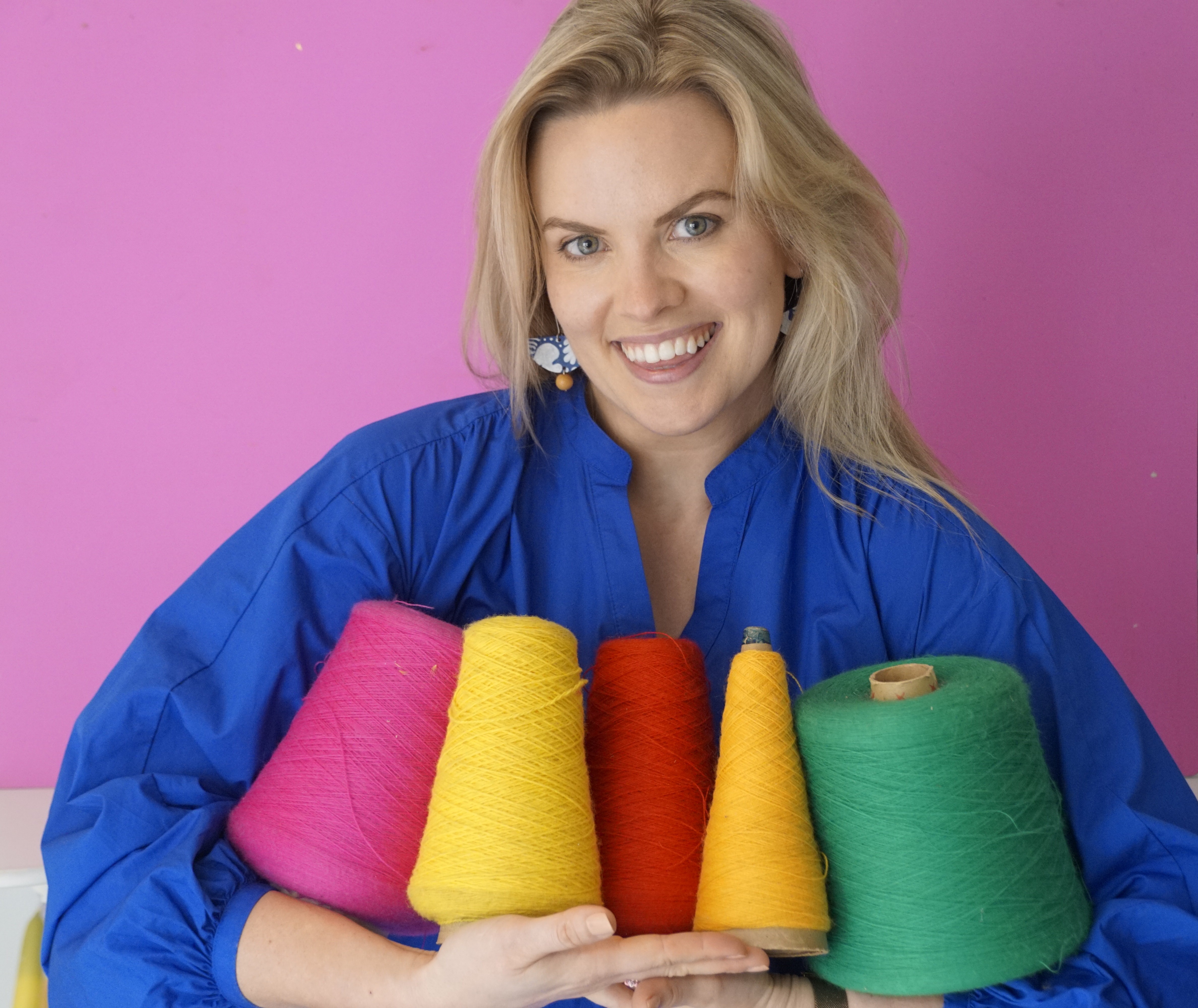 Stacey Barnes smiling and holding colourful cones of yarn against a vivid pink background
