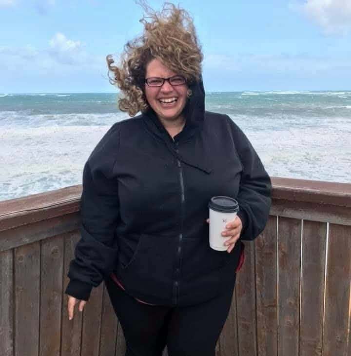 Ronnie laughing at the ocean, hair wild in the wind, holding a coffee